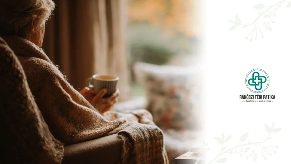 Egy otthonos szobában egy idős ember ül egy fotelben vagy kanapén, takaróba burkolózva. Mellette egy kis asztalon lázcsillapító, egy csésze gőzölgő tea, papírzsebkendő, C-vitamin tabletták és egy orrspray található. A háttérben, az ablakon keresztül halványan látszik az őszi-téli időjárás: lehullott levelek vagy havas táj. A jelenet barátságos, gondoskodó, otthonos és egészségtudatos hangulatot áraszt.